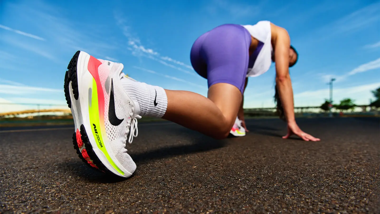 Low-angle action shot of an athlete in purple shorts and a white top crouching in a runner start position, highlighting a modern white running shoe with neon yellow and pink accents against a bright blue sky.