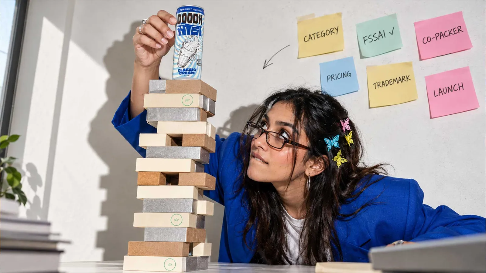 Person balancing a Doodh Soda can on a tower of blocks, with sticky notes for category, FSSAI, co-packer, pricing, trademark and launch on the wall